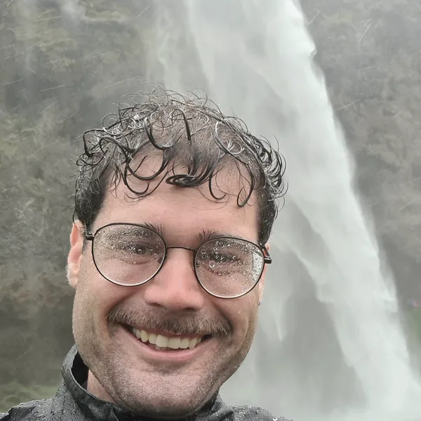 Malte lacht, durchnässt und mit beschlagener Brille, vor einem Wasserfall